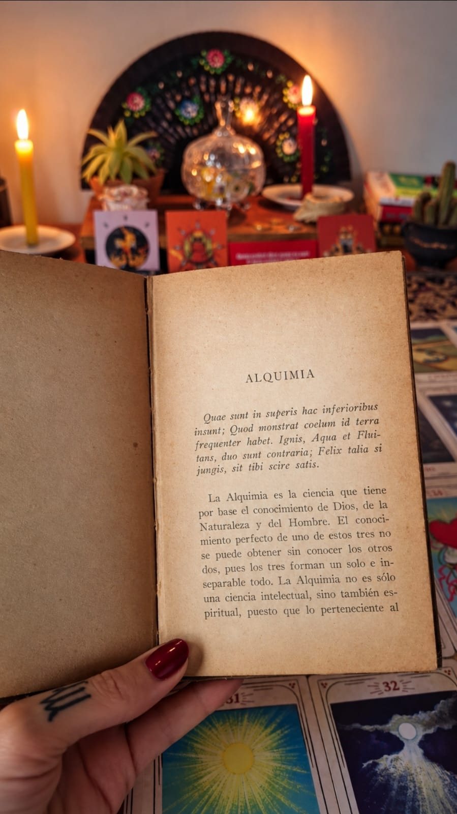 Mão segurando um livro antigo aberto na página Alquimia, diante de um altar com velas acesas e cartas de oráculo sobre a mesa